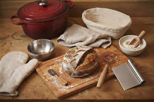 Essential bread baking tools including a Dutch oven, banneton basket, dough whisk, dough scraper, bread lame, and fresh homemade bread on a wooden countertop