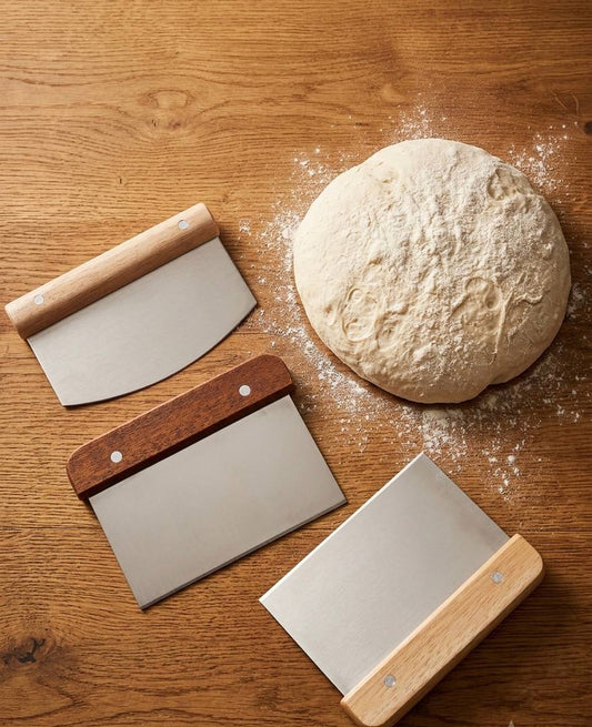 Three dough scrapers on a kitchen countertop beside bread dough on a wooden counter